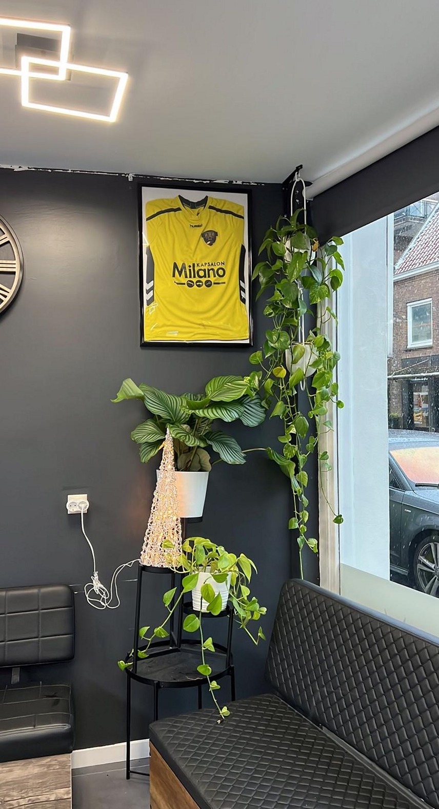 De wachthoek van Kapsalon Milano met planten, een zwart bankje en een ingelijst geel shirt met het Milano-logo
