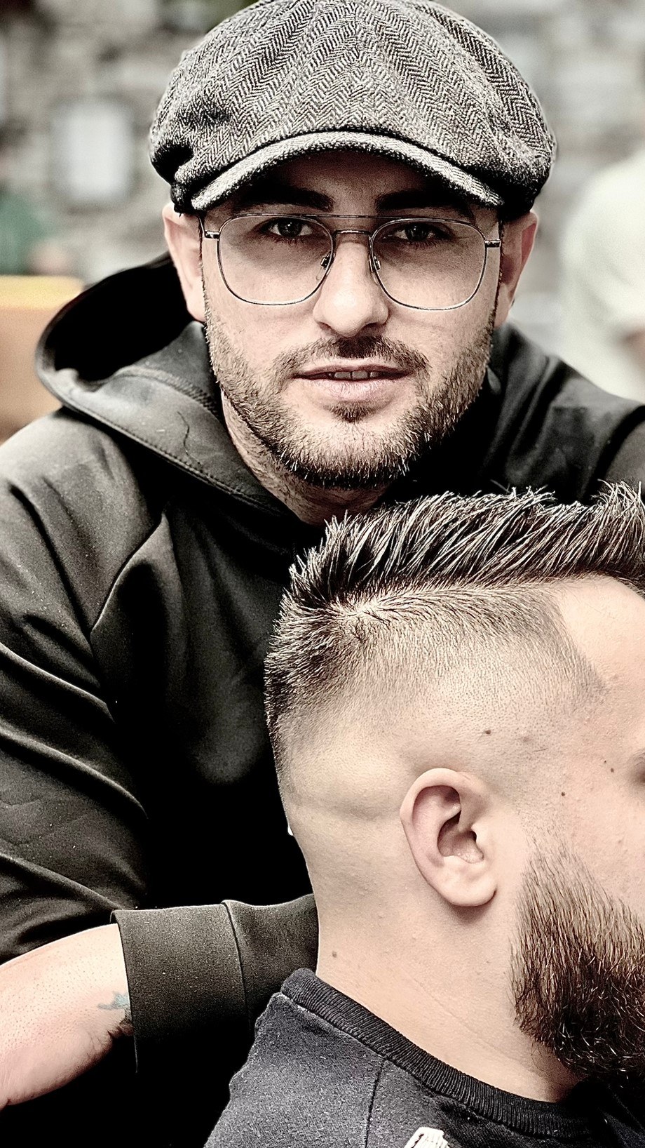 Een van de eigenaren met pet en bril, met voor hem een klant met een vers geknipte fade en baard